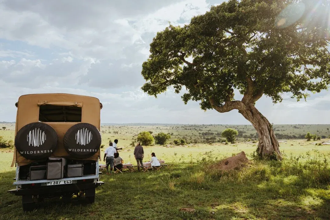 A Wilderness Destinations safari vehicle and guests gathered beneath a broad tree in open grassland